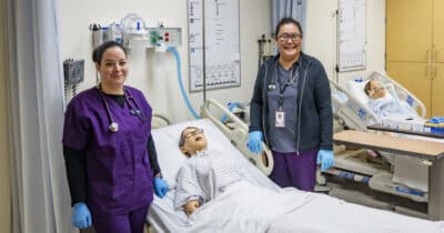 Two licensed practical nursing students pose next to practice mannequin in hospital bed