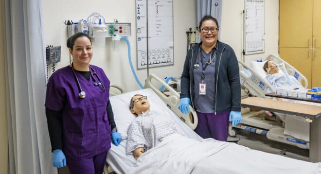 Two licensed practical nursing students pose next to practice mannequin in hospital bed